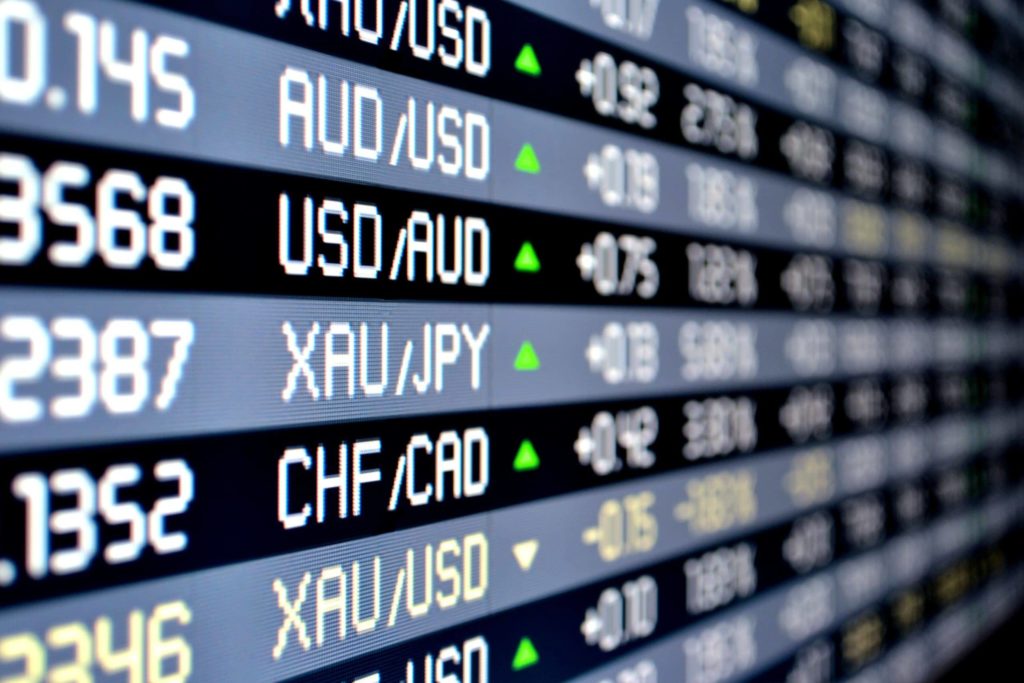 A close-up of an electronic forex board displays various currency pairs and exchange rates, with green arrows indicating positive changes in value.