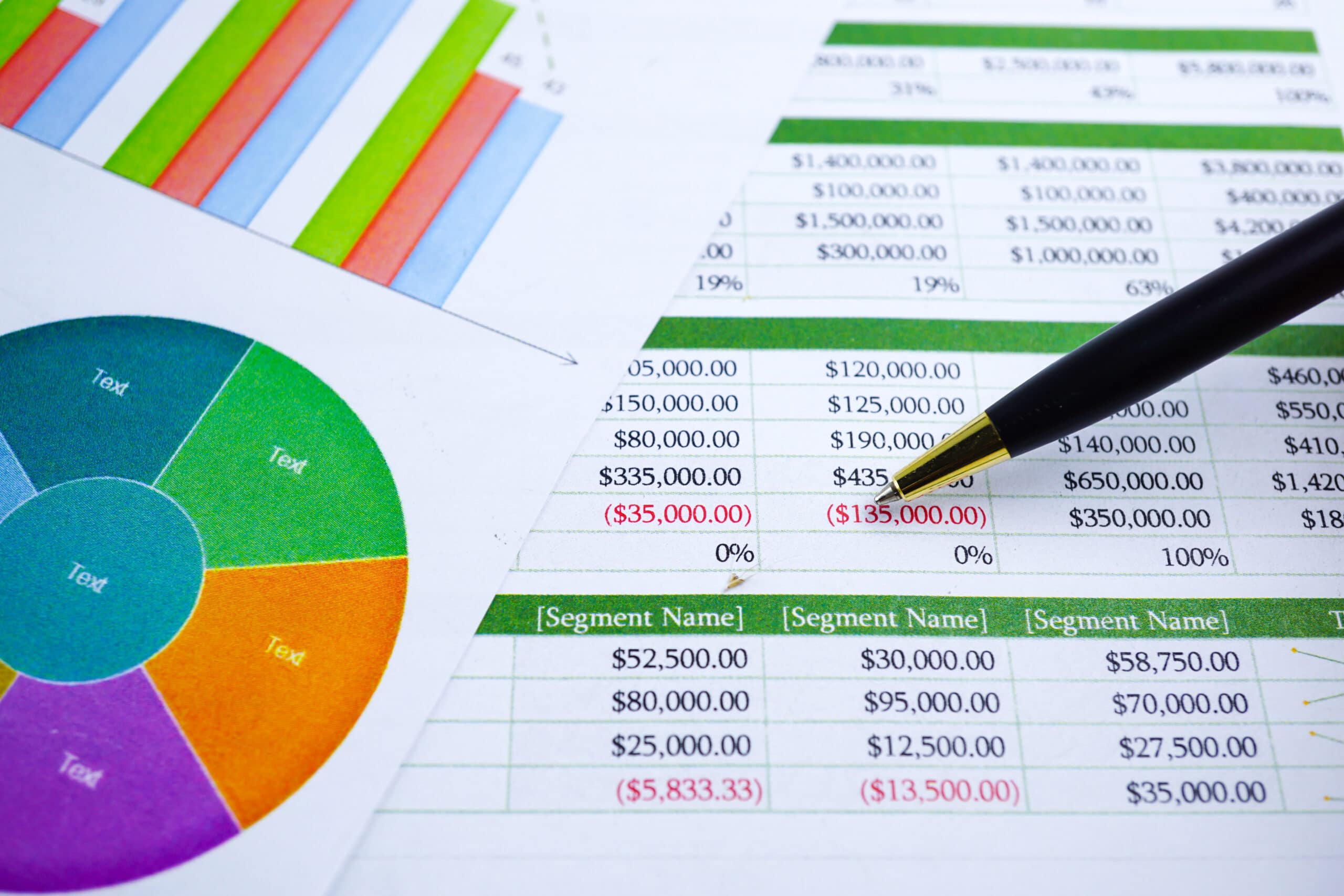 A close-up of financial spreadsheets with charts and graphs. A black pen points to a figure in red, and various numbers and colored segments are visible, indicating financial analysis or business data review.