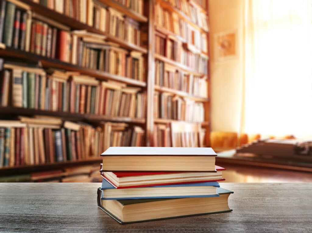 A stack of four books sits on a wooden table in the foreground, with shelves of books filling a sunlit library in the background.