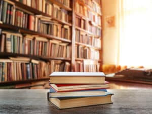 A stack of four books sits on a wooden table in the foreground, with shelves of books filling a sunlit library in the background.