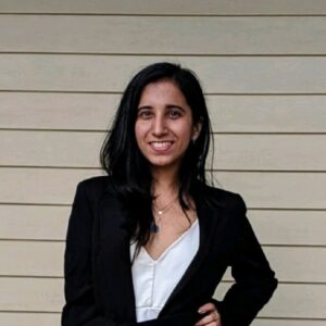 A woman with long dark hair, wearing a black blazer over a white top, stands smiling in front of a light-colored wooden panel wall.