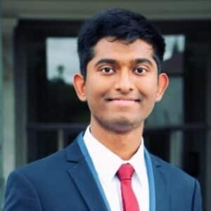 A young man with short black hair, wearing a dark suit, white shirt, and red tie, smiles at the camera. The background is blurred, possibly showing a building entrance.