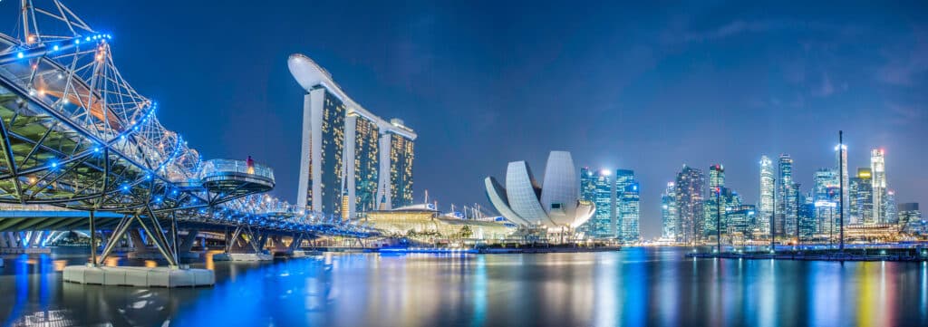 Singapore’s Marina Bay skyline at night, featuring the Helix Bridge on the left, Marina Bay Sands hotel in the center, and illuminated skyscrapers on the right reflected in the calm water.