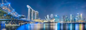 Singapore’s Marina Bay skyline at night, featuring the Helix Bridge on the left, Marina Bay Sands hotel in the center, and illuminated skyscrapers on the right reflected in the calm water.