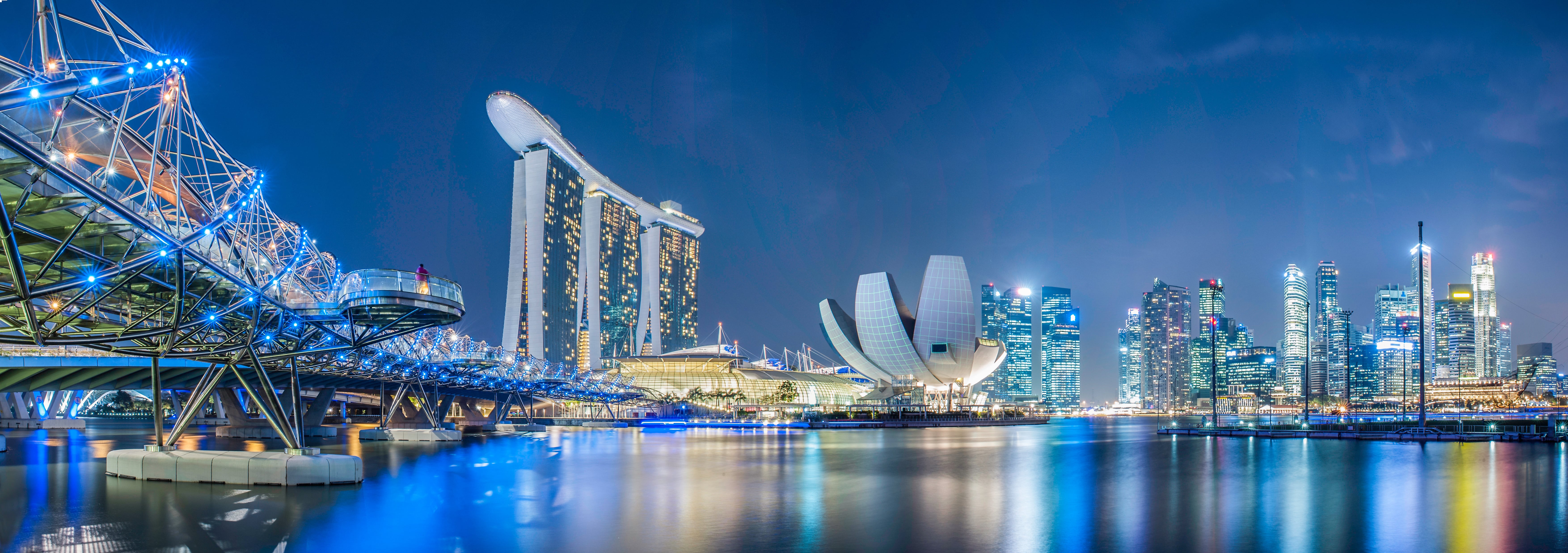 Singapore’s Marina Bay skyline at night, featuring the Helix Bridge on the left, Marina Bay Sands hotel in the center, and illuminated skyscrapers on the right reflected in the calm water.