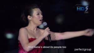 A woman in a sparkly red dress sings passionately on stage, holding a microphone and extending her arm. Subtitles read, That home's about its people too—just like why I moved from Zurich to Singapore. TV channel logos appear in the corner.